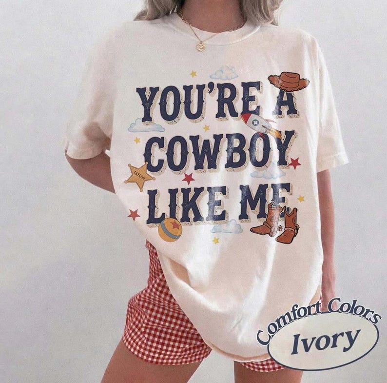 以下が含まれることがあります： アイボリーの半袖Tシャツ。ダークブルーの文字で「YOU'RE A COWBOY LIKE ME」とプリントされ、茶色のカウボーイハット、ロケット、金色の保安官バッジ、カラフルなボール、茶色のカウボーイブーツのグラフィックイラストが周囲に散りばめられています。