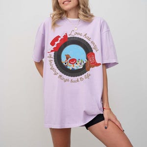 Może przedstawiać: Jasnofioletowy t-shirt z grafiką czarnej opony otaczającej scenę z niebieskim niebem i postaciami z kresk&oacute;wek. Wz&oacute;r zawiera czerwony kapelusz, czerwony kowbojski but oraz napis "Love has ways of bringing things back to life".