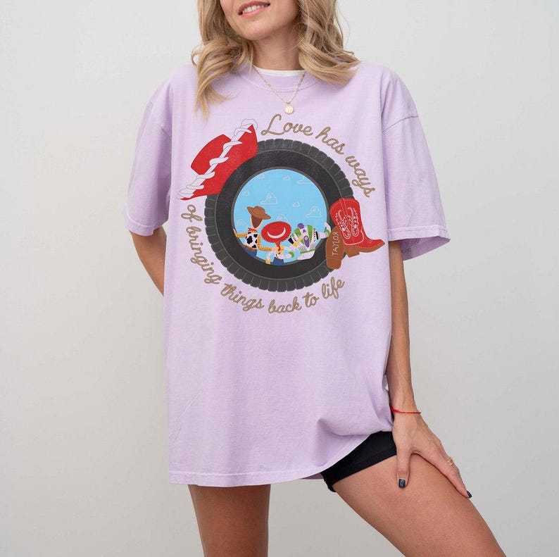 Może przedstawiać: Jasnofioletowy t-shirt z grafiką czarnej opony otaczającej scenę z niebieskim niebem i postaciami z kresk&oacute;wek. Wz&oacute;r zawiera czerwony kapelusz, czerwony kowbojski but oraz napis "Love has ways of bringing things back to life".