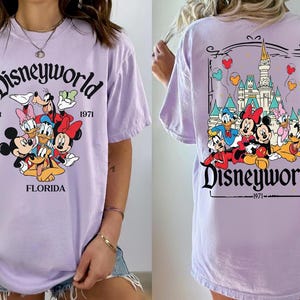 May include: A light purple t-shirt featuring a Disney World graphic. The front of the shirt displays Mickey Mouse, Minnie Mouse, Donald Duck, Daisy Duck, Goofy, and Pluto. The back of the shirt shows the Disney World castle and characters.