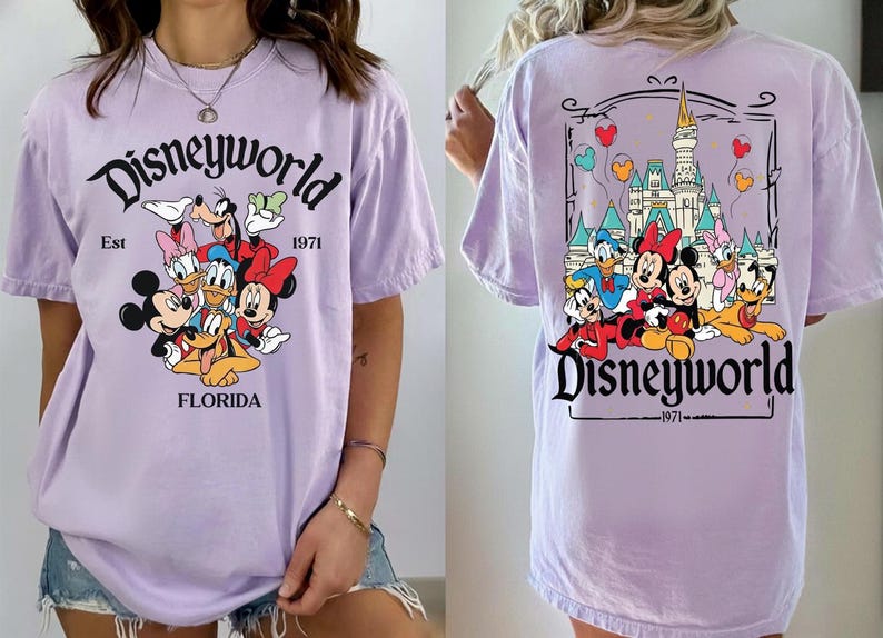 May include: A light purple t-shirt featuring a Disney World graphic. The front of the shirt displays Mickey Mouse, Minnie Mouse, Donald Duck, Daisy Duck, Goofy, and Pluto. The back of the shirt shows the Disney World castle and characters.
