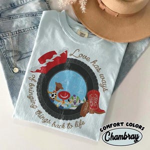 Może przedstawiać: Jasnoniebieski t-shirt z grafiką czarnej opony otaczającej scenę z lalką kowboja i lalką strażnika kosmosu. Wz&oacute;r zawiera czerwony kapelusz kowbojski, czerwony but kowbojski oraz napis Love has ways of bringing things back to life.