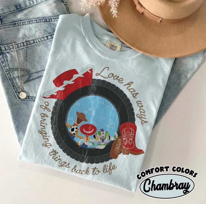Może przedstawiać: Jasnoniebieski t-shirt z grafiką czarnej opony otaczającej scenę z lalką kowboja i lalką strażnika kosmosu. Wz&oacute;r zawiera czerwony kapelusz kowbojski, czerwony but kowbojski oraz napis Love has ways of bringing things back to life.