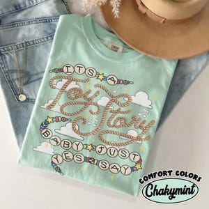 May include: A light mint green t-shirt featuring a graphic design with rope-style lettering that reads "ITS A Toy Story BABY JUST YES SAY". The design includes colorful beads, small stars, musical notes, and white clouds on a soft fabric.