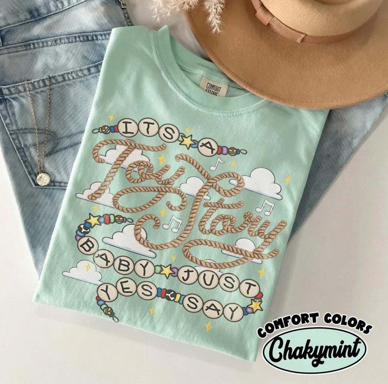 May include: A light mint green t-shirt featuring a graphic design with rope-style lettering that reads "ITS A Toy Story BABY JUST YES SAY". The design includes colorful beads, small stars, musical notes, and white clouds on a soft fabric.