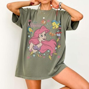 May include: Olive green t-shirt featuring a graphic of Ariel from Disney's The Little Mermaid. The design includes Ariel with pink hair, a purple seashell bra, and various sea creatures. The shirt has short sleeves and a relaxed fit.
