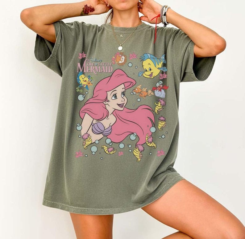 May include: Olive green t-shirt featuring a graphic of Ariel from Disney's The Little Mermaid. The design includes Ariel with pink hair, a purple seashell bra, and various sea creatures. The shirt has short sleeves and a relaxed fit.