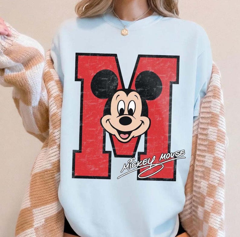 May include: Light blue sweatshirt featuring a distressed red letter "M" with a Mickey Mouse graphic. The design includes the words "Mickey Mouse" in a cursive font. The sweatshirt is a casual style.