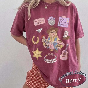 May include: A berry-colored short-sleeve t-shirt featuring a graphic print of a cowgirl, a cowboy doll, a space ranger doll, a disco ball, a guitar, a horseshoe, a sheriff star, a beaded bracelet, and text reading Toy Box Tour 2026.