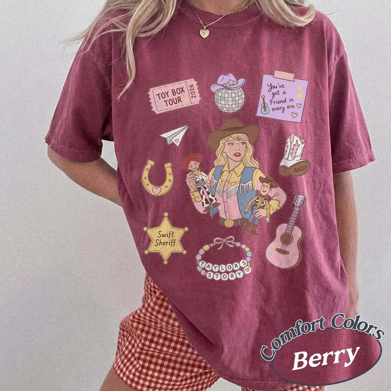 May include: A berry-colored short-sleeve t-shirt featuring a graphic print of a cowgirl, a cowboy doll, a space ranger doll, a disco ball, a guitar, a horseshoe, a sheriff star, a beaded bracelet, and text reading Toy Box Tour 2026.
