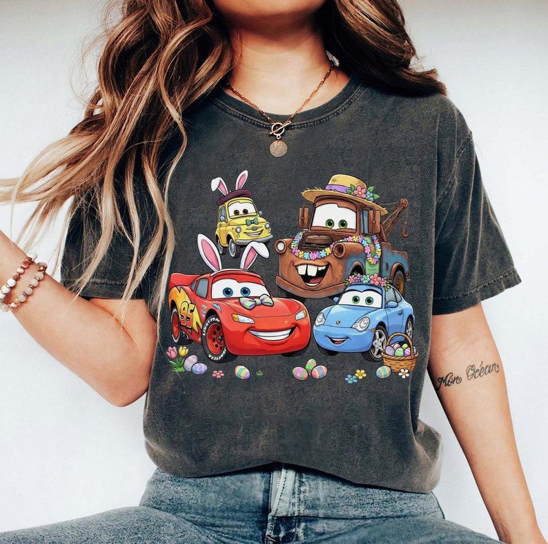 Pode incluir: T-shirt cinzento escuro com um design de P&aacute;scoa com carros de desenho animado. O design inclui um carro de corrida vermelho, um carro azul, um carro amarelo e um cami&atilde;o de reboque castanho, todos com decora&ccedil;&otilde;es de P&aacute;scoa e ovos de P&aacute;scoa.