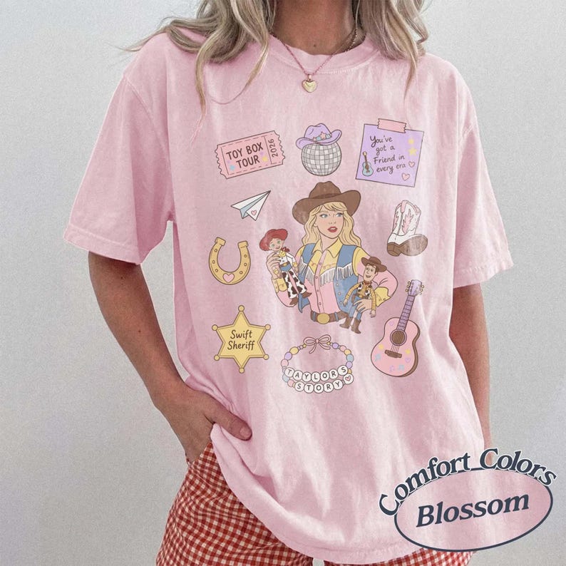 May include: A light pink short-sleeve t-shirt featuring a graphic print of a cowgirl, a guitar, a horseshoe, a sheriff star labeled Swift Sheriff, a disco ball, a paper airplane, a cowboy boot, and text reading Toy Box Tour 2026.