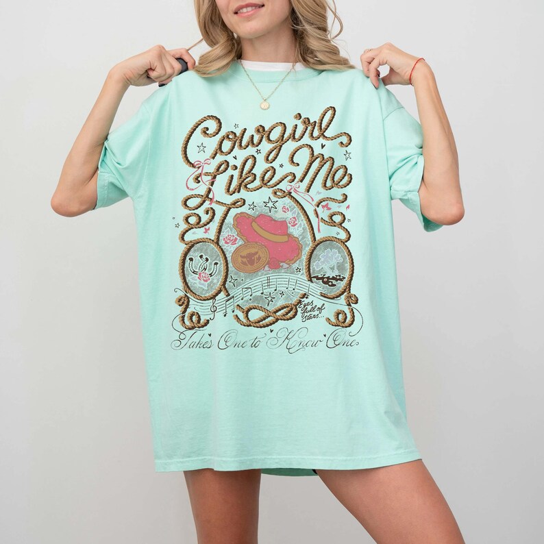 May include: A light mint green t-shirt featuring a graphic print with the text Cowgirl Like Me, Eyes full of Stars, and Takes One to Know One. The design includes a rope border, a pink cowboy hat, a horseshoe, and musical staff lines.