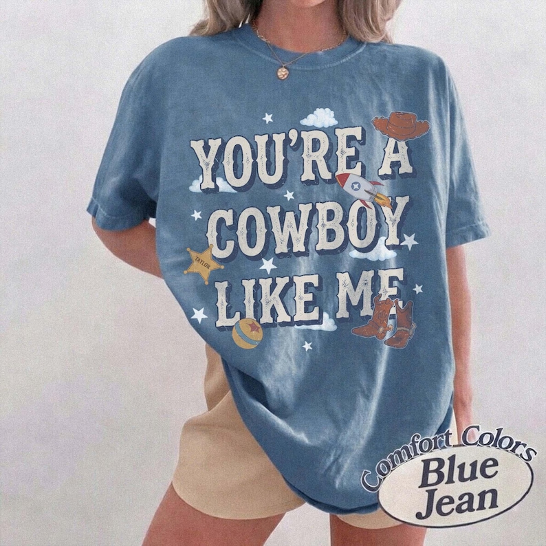 以下が含まれることがあります： 白い文字で「YOU'RE A COWBOY LIKE ME」と書かれた青い半袖Tシャツ。グラフィックには、茶色のカウボーイハット、ロケット、黄色い星、ボール、白い雲のイラストが青い背景に描かれています。
