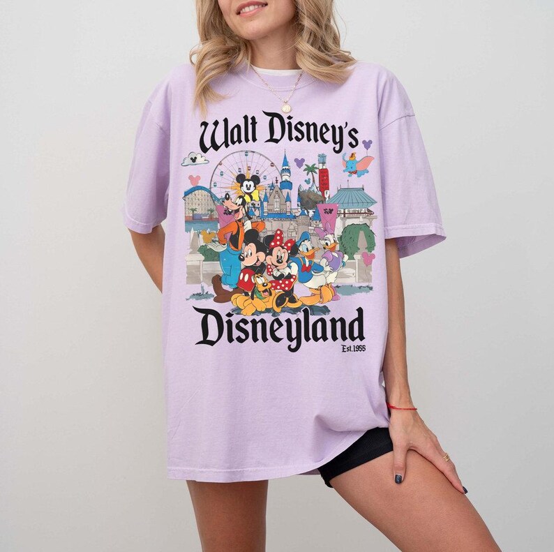 Puede incluir: Camiseta lila claro con un gr&aacute;fico colorido de personajes de Disney y el texto "Walt Disney's Disneyland" en negro. El dise&ntilde;o incluye Mickey Mouse, Minnie Mouse, Donald Duck y otros personajes.