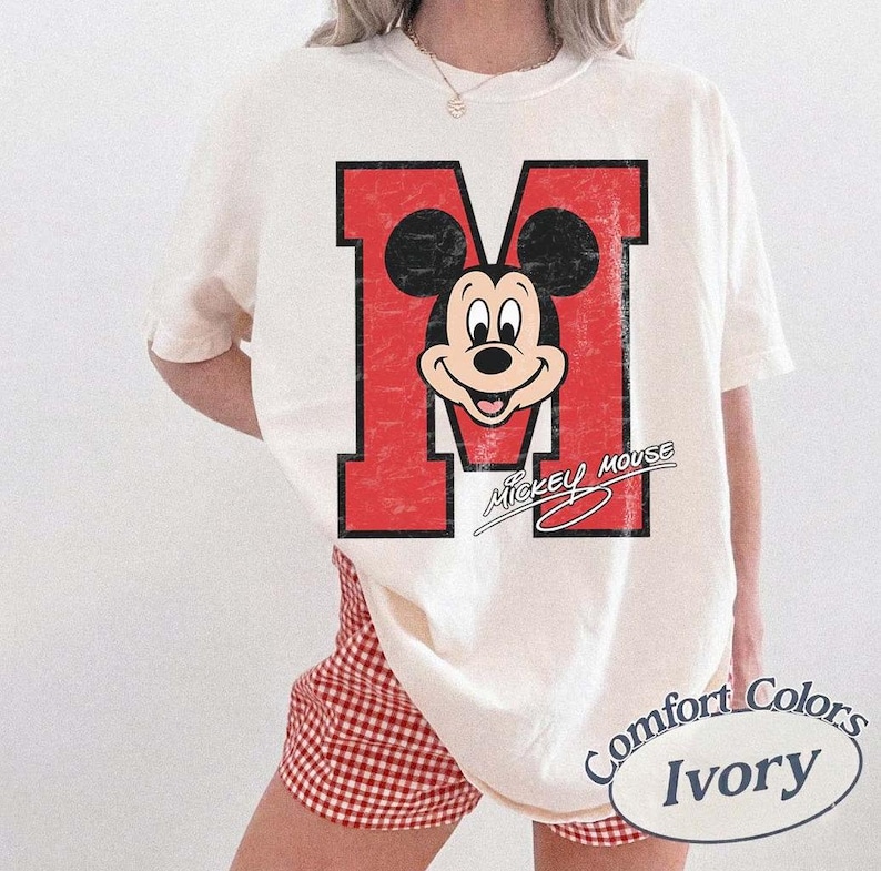May include: Ivory-colored t-shirt featuring a distressed red letter "M" with a Mickey Mouse graphic. The shirt has the words "Mickey Mouse" in a script font below the graphic. The shirt is part of the Comfort Colors collection.