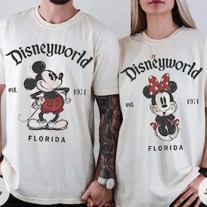 May include: Ivory-colored t-shirts featuring Mickey and Minnie Mouse. The shirts have the text "Disneyworld" above the characters, with "est. 1971" and "Florida" below. Mickey is in red shorts, and Minnie has a red bow.