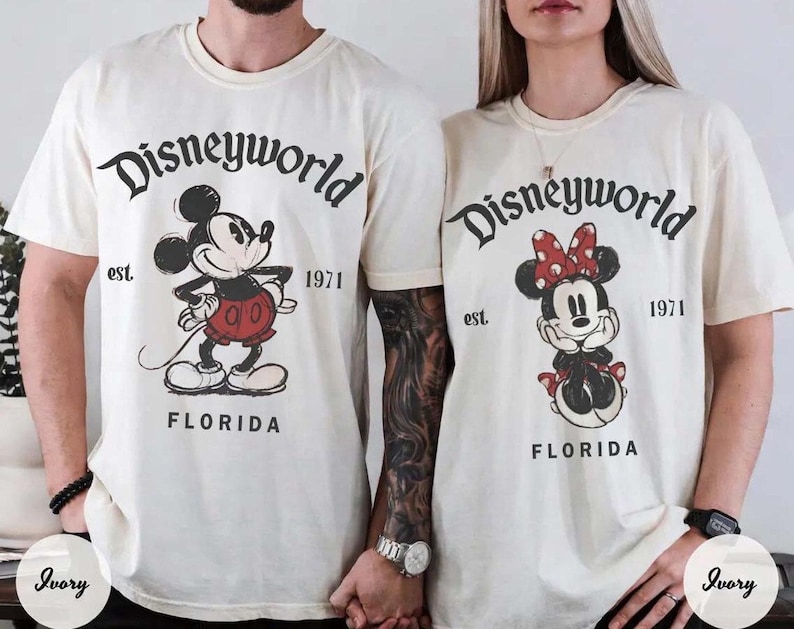 May include: Ivory-colored t-shirts featuring Mickey and Minnie Mouse. The shirts have the text "Disneyworld" above the characters, with "est. 1971" and "Florida" below. Mickey is in red shorts, and Minnie has a red bow.