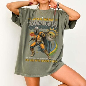 May include: An olive green t-shirt featuring a graphic of The Mandalorian and Grogu. The text reads STAR WARS THE MANDALORIAN, BOUNTY HUNTER, and WHEN YOU CHOOSE TO WALK THE WAY OF THE MANDALORIAN YOU ARE BOTH HUNTER & PREY in yellow and white.