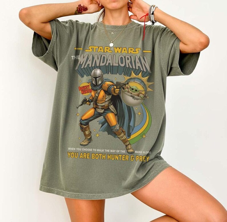 May include: An olive green t-shirt featuring a graphic of The Mandalorian and Grogu. The text reads STAR WARS THE MANDALORIAN, BOUNTY HUNTER, and WHEN YOU CHOOSE TO WALK THE WAY OF THE MANDALORIAN YOU ARE BOTH HUNTER & PREY in yellow and white.