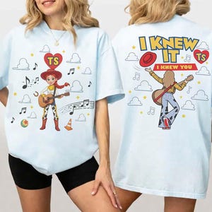 Puede incluir: Una camiseta azul claro con un gr&aacute;fico de una vaquera tocando la guitarra en la parte delantera, y un dise&ntilde;o trasero con el texto I KNEW IT I KNEW YOU, un sombrero rojo, una guitarra y notas musicales rodeadas de nubes y estrellas amarillas.