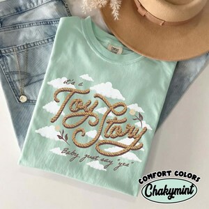 May include: A light mint green t-shirt featuring a graphic of white clouds and brown rope-textured lettering that reads "it's a Toy Story" and "Baby, just say "yes"" with small floral accents. The shirt has a crew neckline and a tag.