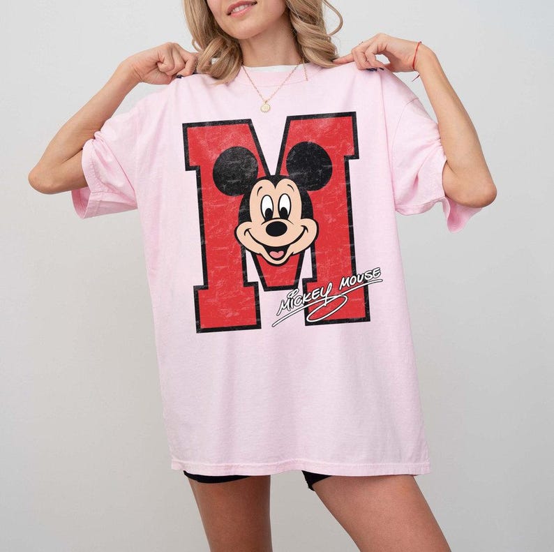 May include: A light pink t-shirt featuring a large red letter "M" with a distressed texture. The letter incorporates a black and white Mickey Mouse graphic. The words "Mickey Mouse" are printed below the graphic in a cursive font.