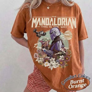 May include: Burnt orange t-shirt featuring a graphic of The Mandalorian and Grogu surrounded by flowers. The text "THE MANDALORIAN - THIS IS THE WAY" is printed across the top. The shirt has a Comfort Colors label.