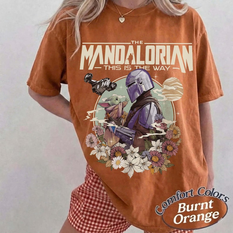 May include: Burnt orange t-shirt featuring a graphic of The Mandalorian and Grogu surrounded by flowers. The text "THE MANDALORIAN - THIS IS THE WAY" is printed across the top. The shirt has a Comfort Colors label.