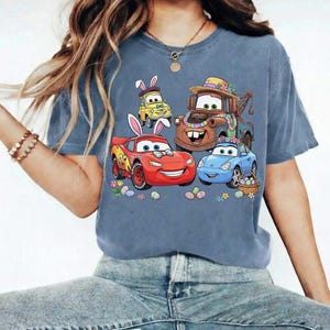 Pode incluir: Uma camiseta azul com um gr&aacute;fico colorido de carros animados decorados com orelhas de coelho da P&aacute;scoa e detalhes florais. Os carros s&atilde;o cercados por ovos de P&aacute;scoa e flores, criando um design festivo.