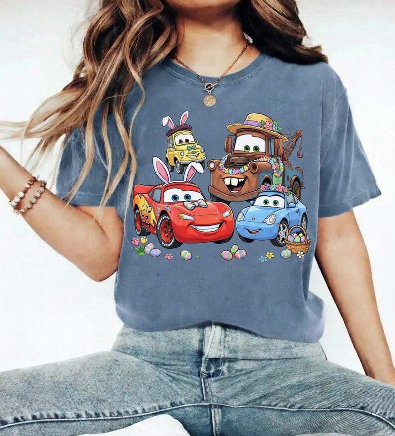 Pode incluir: Uma camiseta azul com um gr&aacute;fico colorido de carros animados decorados com orelhas de coelho da P&aacute;scoa e detalhes florais. Os carros s&atilde;o cercados por ovos de P&aacute;scoa e flores, criando um design festivo.