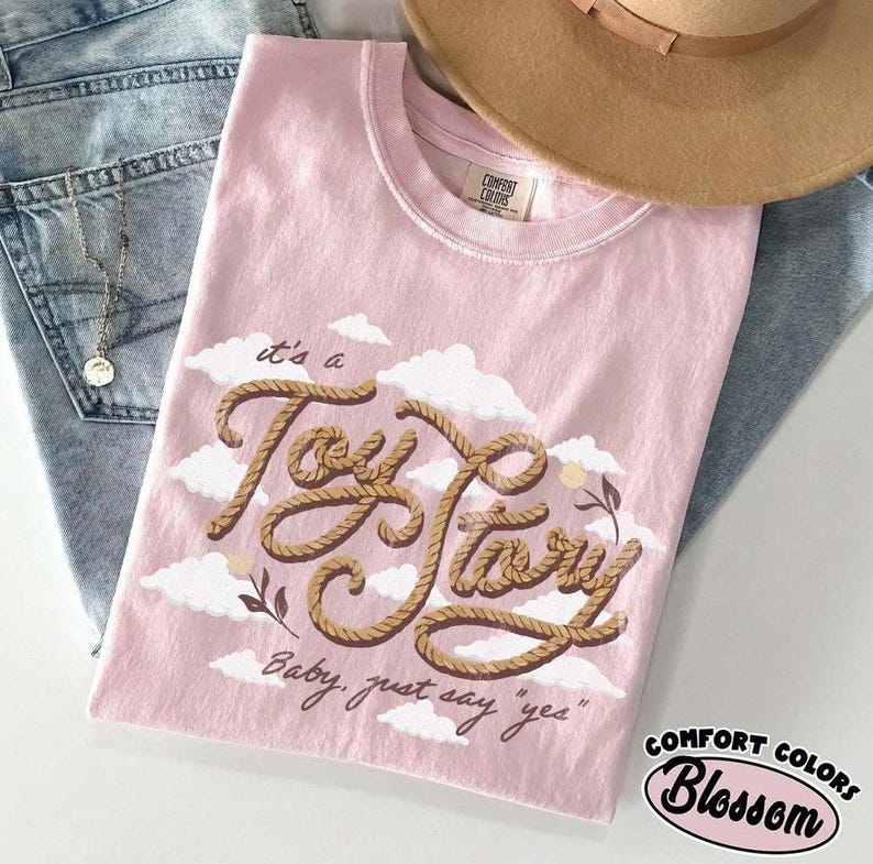 May include: A light pink t-shirt featuring a graphic of the text "it's a Toy Story" written in a brown rope-like font, surrounded by white clouds and small flowers. Below the main text, the phrase "Baby, just say 'yes'" is printed in cursive.