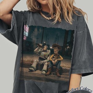 Puede incluir: Camiseta Comfort Colors gris oscuro con un gr&aacute;fico de Buzz Lightyear y Woody de Toy Story sentados en una acera, bebiendo. La imagen tiene un aspecto vintage y desgastado. La camiseta tiene cuello redondo y mangas cortas.