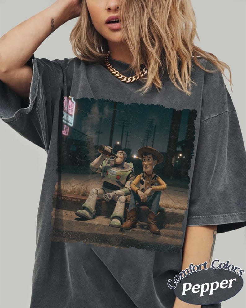 Puede incluir: Camiseta Comfort Colors gris oscuro con un gr&aacute;fico de Buzz Lightyear y Woody de Toy Story sentados en una acera, bebiendo. La imagen tiene un aspecto vintage y desgastado. La camiseta tiene cuello redondo y mangas cortas.
