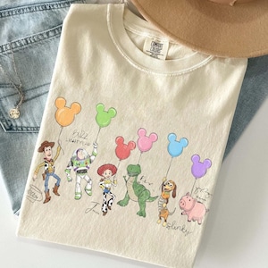 Puede incluir: Camiseta color crema con una colorida ilustraci&oacute;n de personajes de Toy Story sosteniendo globos con forma de Mickey Mouse. Los personajes incluyen a Woody, Buzz Lightyear, Jessie, Rex, Slinky Dog y Hamm. La camiseta es de material suave.