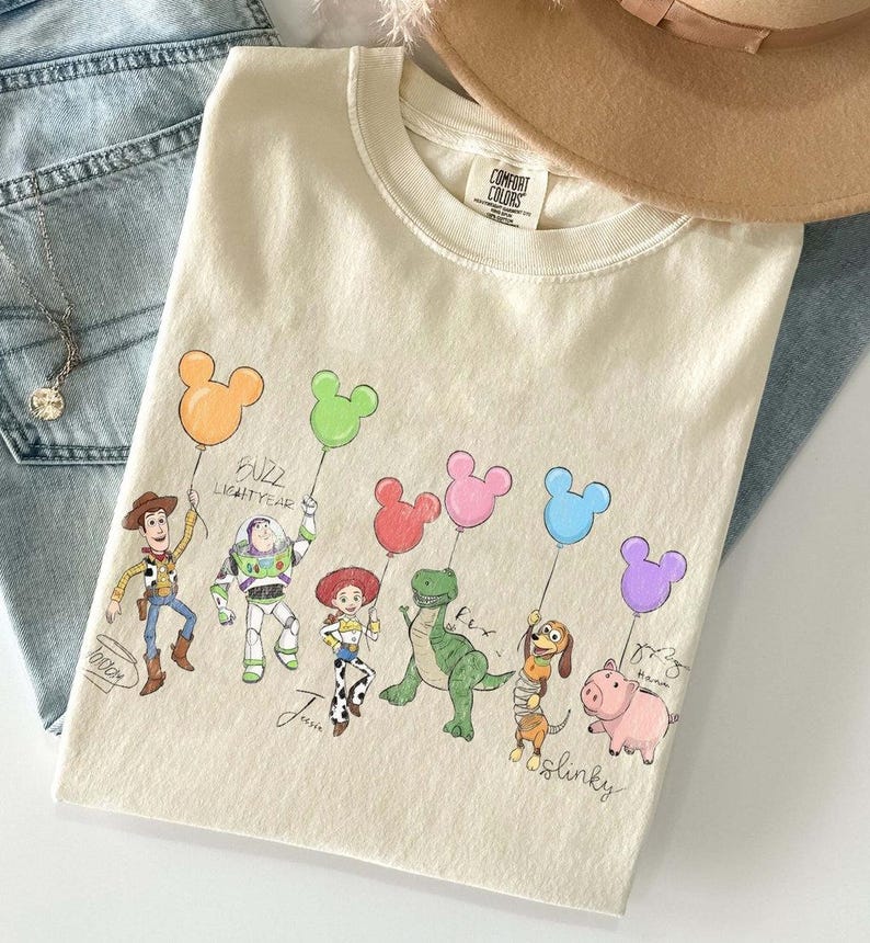 Puede incluir: Camiseta color crema con una colorida ilustraci&oacute;n de personajes de Toy Story sosteniendo globos con forma de Mickey Mouse. Los personajes incluyen a Woody, Buzz Lightyear, Jessie, Rex, Slinky Dog y Hamm. La camiseta es de material suave.