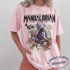 May include: Pink t-shirt featuring a graphic of The Mandalorian and Grogu, with the text "THE MANDALORIAN - THIS IS THE WAY." The design includes a floral arrangement and a planet. The shirt is from Comfort Colors.