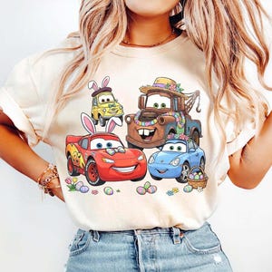 Pode incluir: Camiseta bege com um gr&aacute;fico colorido de carros animados decorados para a P&aacute;scoa. Os carros incluem um carro de corrida vermelho, um carro esportivo azul, um carro amarelo e um caminh&atilde;o de reboque marrom, todos com decora&ccedil;&otilde;es de P&aacute;scoa. Ovos de P&aacute;scoa e flores tamb&eacute;m est&atilde;o presentes.