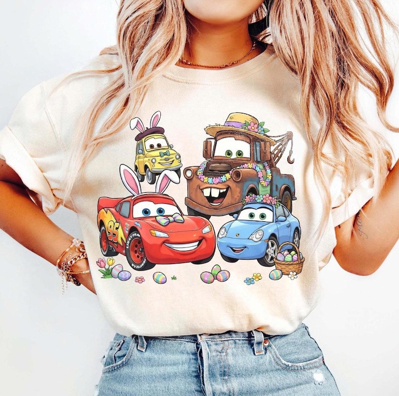 Pode incluir: Camiseta bege com um gr&aacute;fico colorido de carros animados decorados para a P&aacute;scoa. Os carros incluem um carro de corrida vermelho, um carro esportivo azul, um carro amarelo e um caminh&atilde;o de reboque marrom, todos com decora&ccedil;&otilde;es de P&aacute;scoa. Ovos de P&aacute;scoa e flores tamb&eacute;m est&atilde;o presentes.