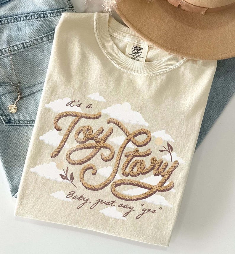 May include: A cream-colored t-shirt featuring a graphic of the text "it's a Toy Story" written in a brown rope-like font, accompanied by the text "Baby, just say 'yes'" and white cloud illustrations. The shirt has a Comfort Colors brand label.