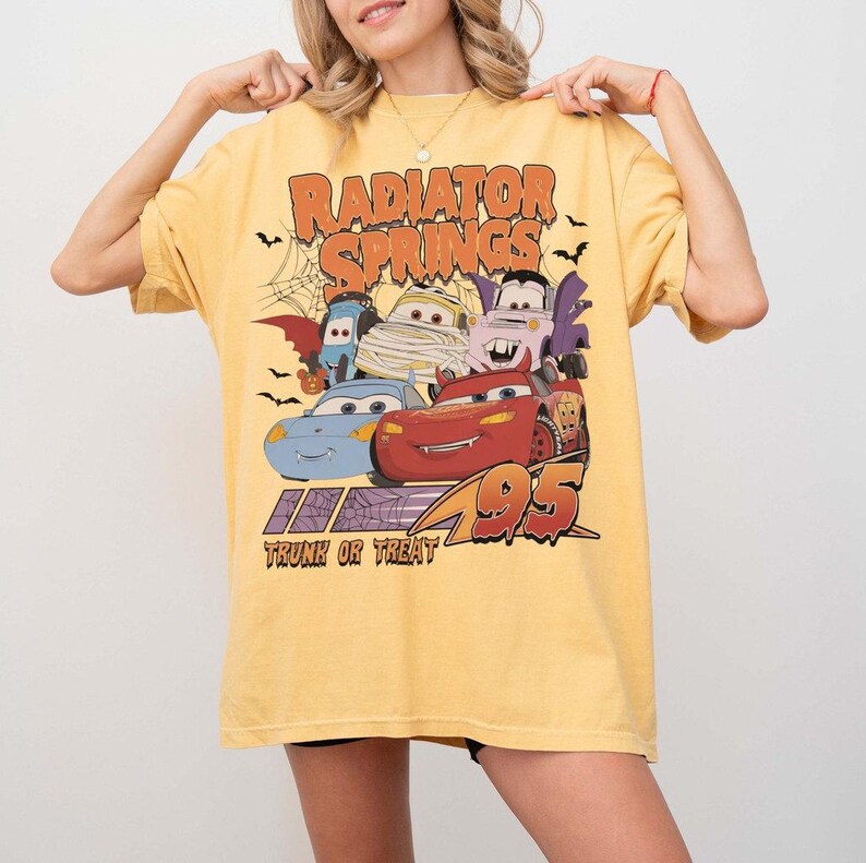May include: Yellow t-shirt featuring a graphic of characters from the movie Cars, dressed in Halloween costumes. The text "Radiator Springs" and "Trunk or Treat" are also displayed on the shirt.