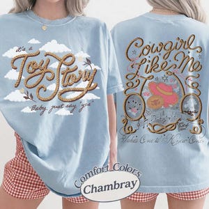 May include: A light blue t-shirt featuring a front graphic with white clouds and the text "it's a Toy Story Baby, just say "yes"" in a rope-style font. The back graphic displays "Cowgirl Like Me" with a pink hat, rope, and musical notes.
