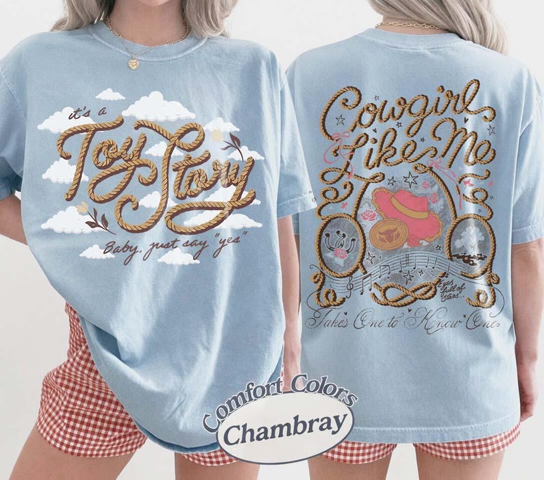 May include: A light blue t-shirt featuring a front graphic with white clouds and the text "it's a Toy Story Baby, just say "yes"" in a rope-style font. The back graphic displays "Cowgirl Like Me" with a pink hat, rope, and musical notes.