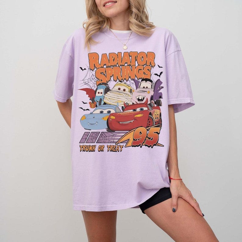 May include: A light purple t-shirt featuring a graphic of characters from the movie Cars, with the text "Radiator Springs" and "Trunk or Treat 95". The design includes cartoon cars and Halloween-themed elements.