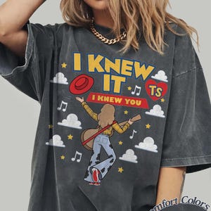 May include: A charcoal gray oversized t-shirt featuring a graphic print with the text I KNEW IT and I KNEW YOU. The design includes a person playing a guitar, a red hat, a red heart with TS, musical notes, stars, and white clouds on the front.