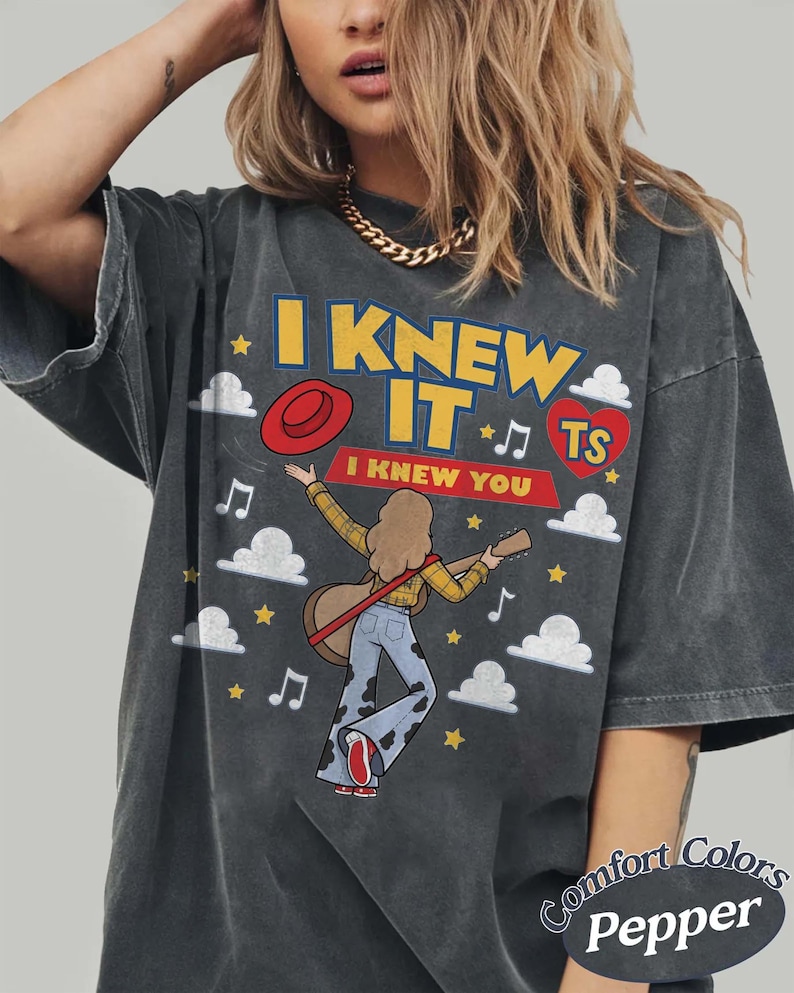 May include: A charcoal gray oversized t-shirt featuring a graphic print with the text I KNEW IT and I KNEW YOU. The design includes a person playing a guitar, a red hat, a red heart with TS, musical notes, stars, and white clouds on the front.