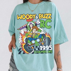 May include: A light blue t-shirt featuring a graphic of Woody, Buzz Lightyear, and RC from Toy Story. The design includes the text "WOODY & BUZZ & RC 1995 CHAMPIONS" with flames and a checkered flag.
