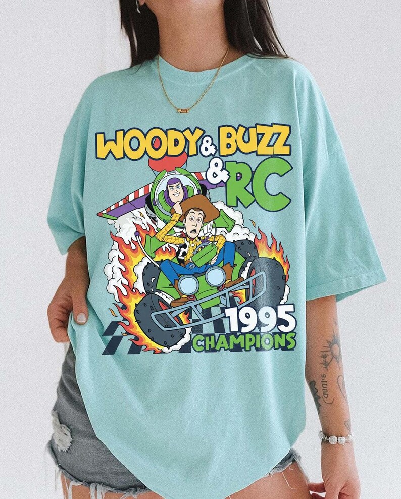 May include: A light blue t-shirt featuring a graphic of Woody, Buzz Lightyear, and RC from Toy Story. The design includes the text "WOODY & BUZZ & RC 1995 CHAMPIONS" with flames and a checkered flag.