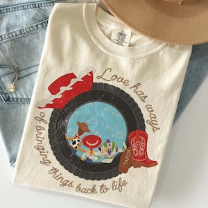 Może przedstawiać: Kremowy t-shirt z grafiką czarnej opony otaczającej błękitne niebo z chmurami i trzema postaciami z kresk&oacute;wek. Wz&oacute;r zawiera czerwony kapelusz kowbojski, czerwony kowbojski but oraz napis Love has ways of bringing things back to life.