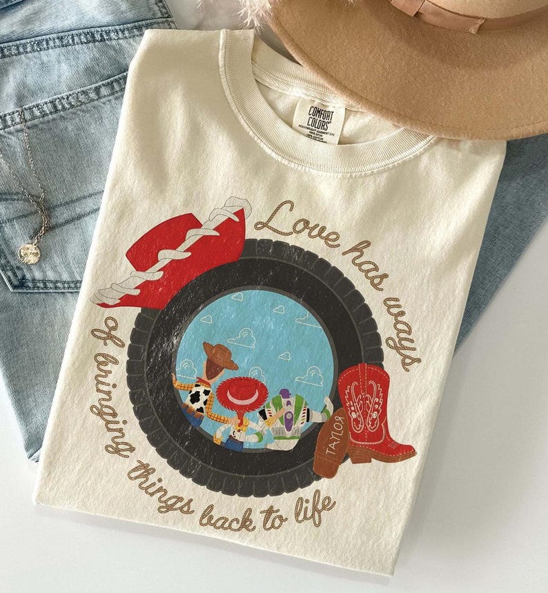 Może przedstawiać: Kremowy t-shirt z grafiką czarnej opony otaczającej błękitne niebo z chmurami i trzema postaciami z kresk&oacute;wek. Wz&oacute;r zawiera czerwony kapelusz kowbojski, czerwony kowbojski but oraz napis Love has ways of bringing things back to life.